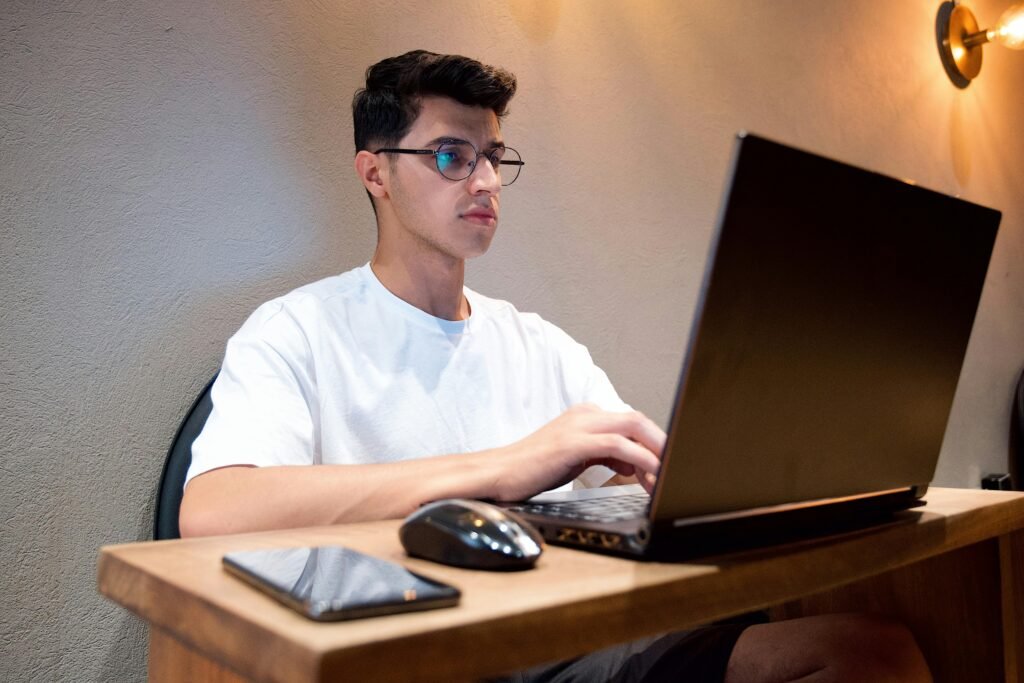 Man in white shirt using laptop at desk indoors with smartphone and mouse nearby.