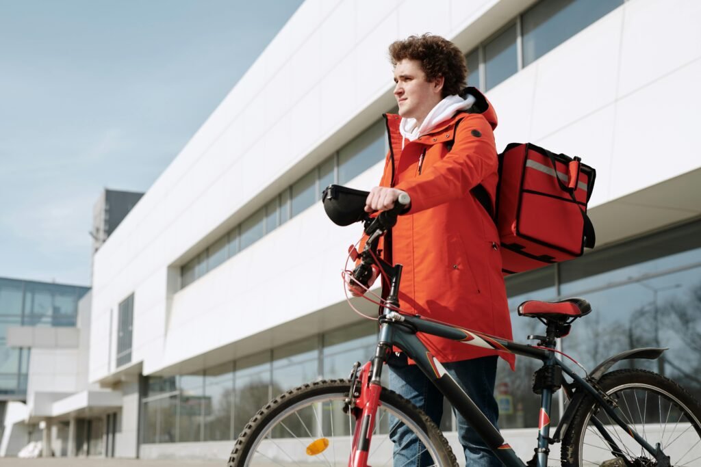 Man with bicycle and delivery backpack in urban setting, vibrant orange coat, modern architecture background.